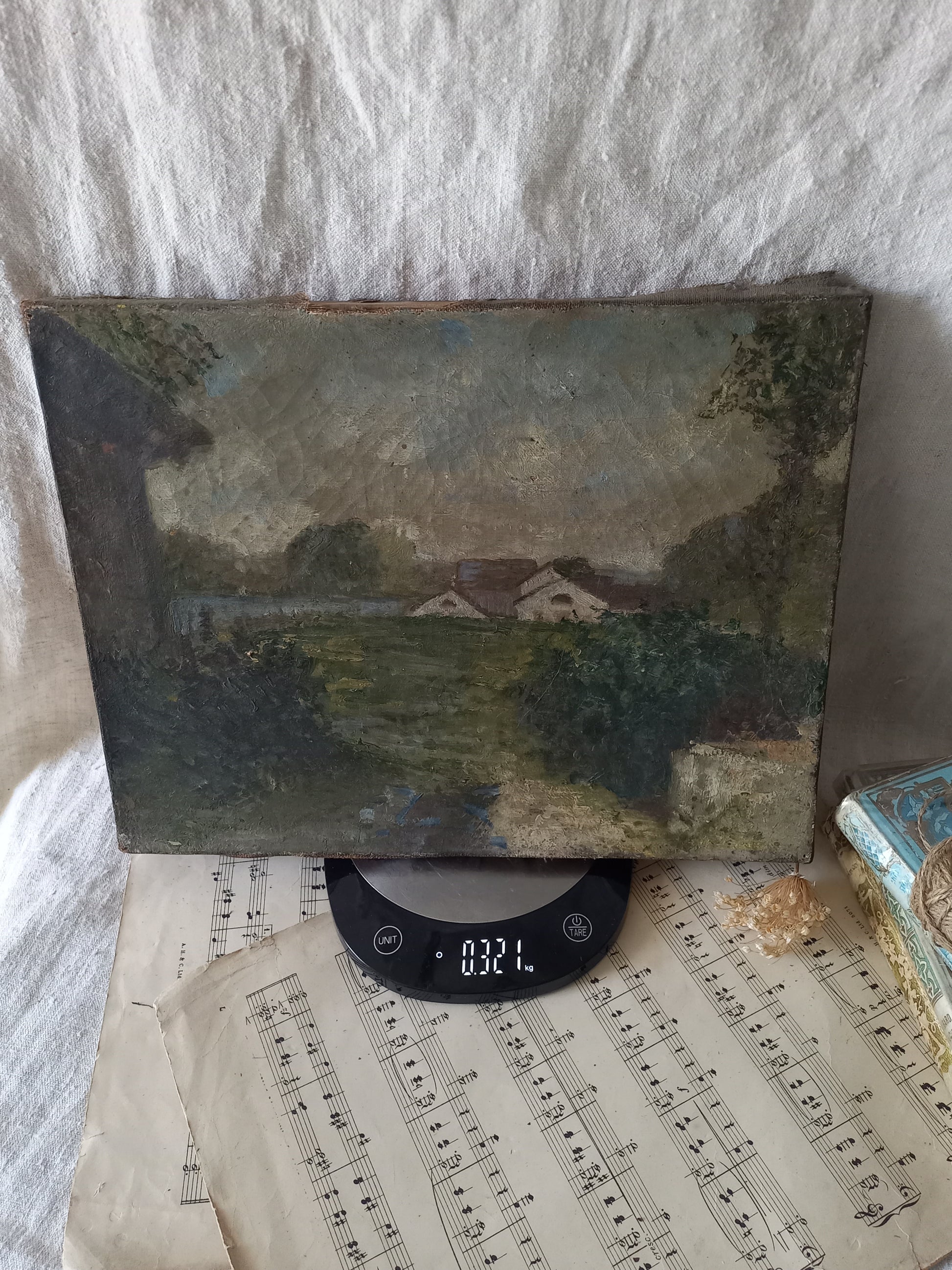 An antique oil landscape painting on canvas, displayed on a digital scale over vintage sheet music against a draped white fabric background.