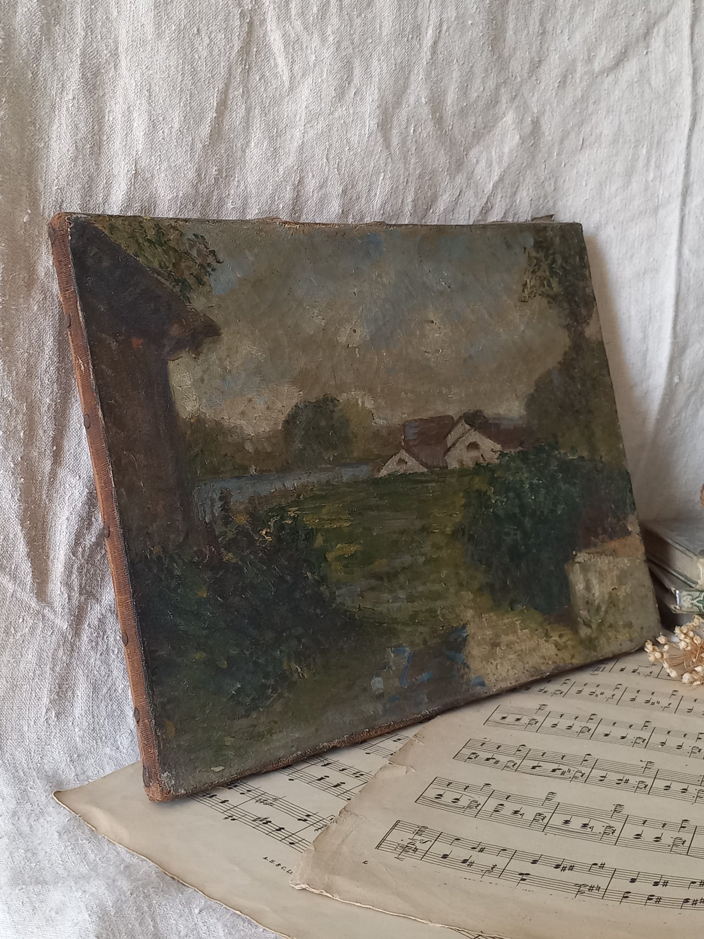 An antique oil landscape painting on canvas, depicting a rural scene with houses, trees, and a path, displayed on top of vintage sheet music against a draped white fabric background.