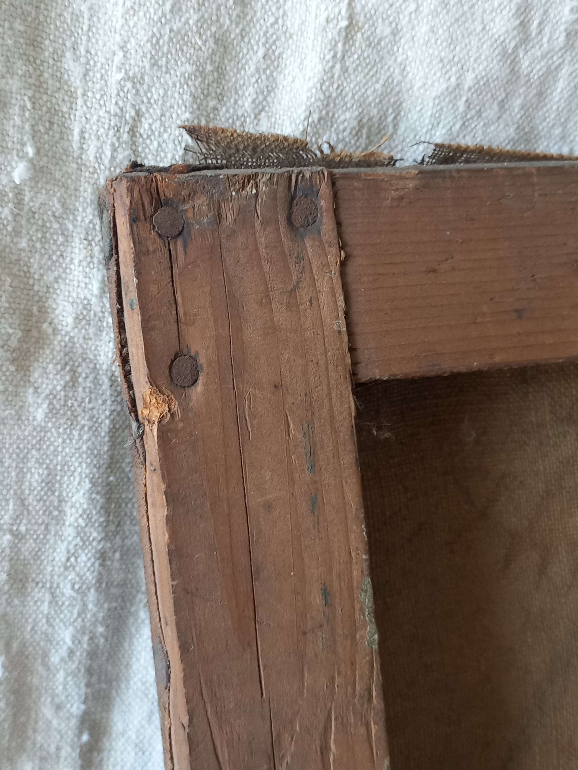 Close-up of a rustic wooden frame corner with weathered brown wood, visible nail heads, and a textured off-white fabric background.