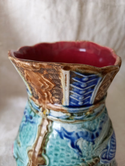 Close-up of an antique majolica pitcher with a rich glaze set against a draped white fabric background.