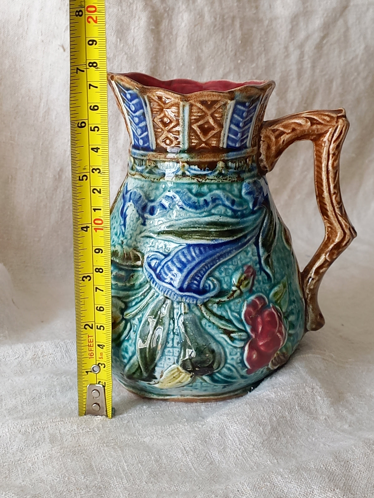 An antique majolica pitcher with a rich glaze set against a draped white fabric background shown next to a yellow tape measure for reference.