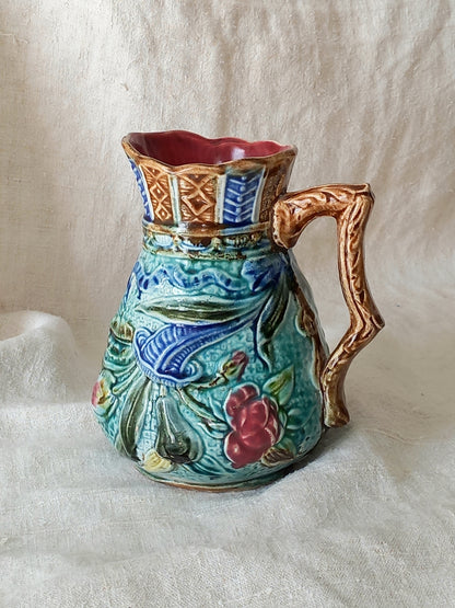 An antique majolica pitcher with a rich glaze set against a draped white fabric background.