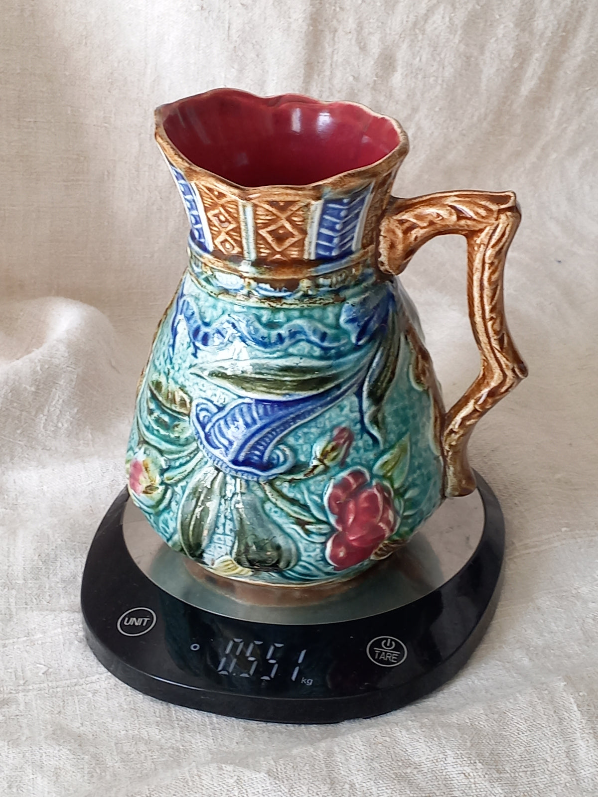 An antique majolica pitcher with a rich glaze set against a draped white fabric background.