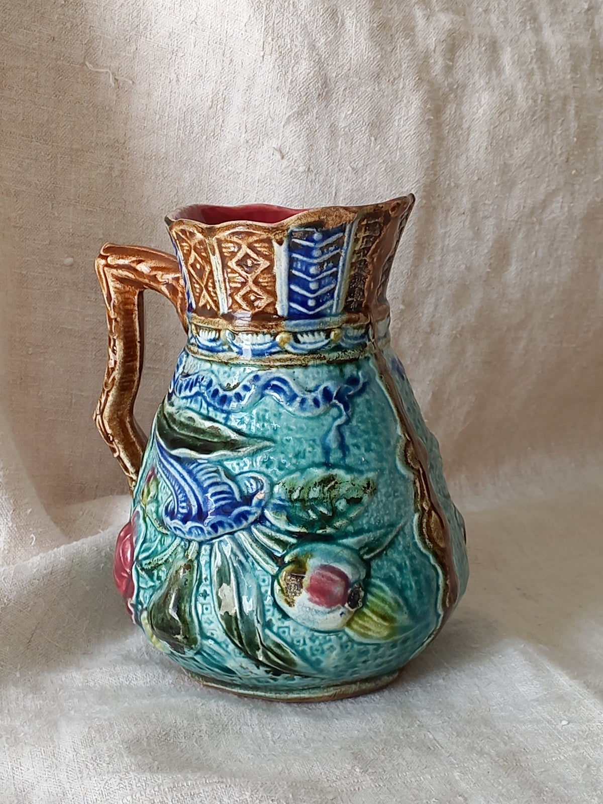 An antique majolica pitcher with a rich glaze set against a draped white fabric background.