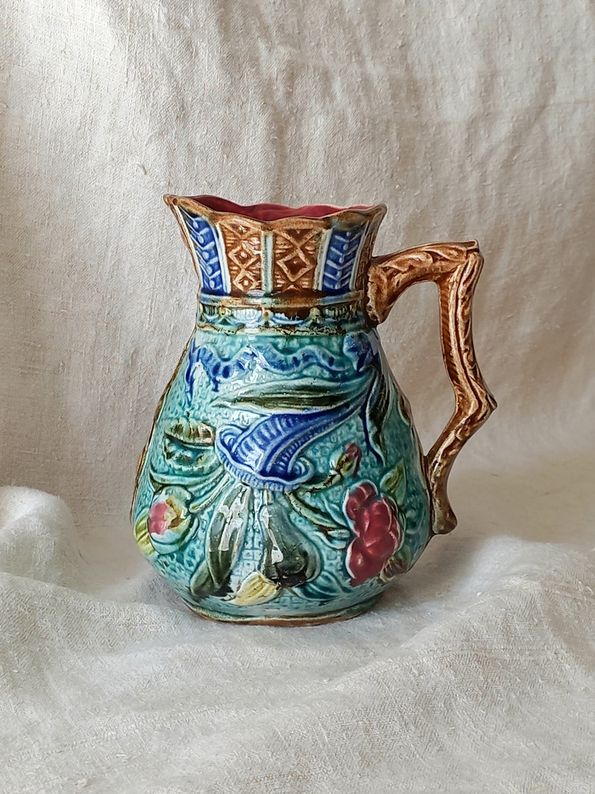 An antique majolica pitcher with a rich glaze set against a draped white fabric background.