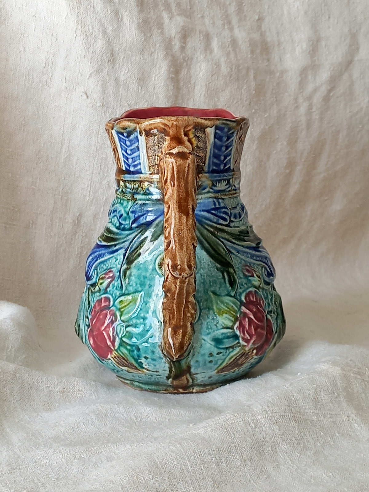 An antique majolica pitcher with a rich glaze set against a draped white fabric background.