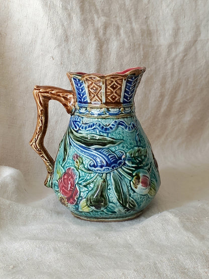 An antique majolica pitcher with a rich glaze set against a draped white fabric background.