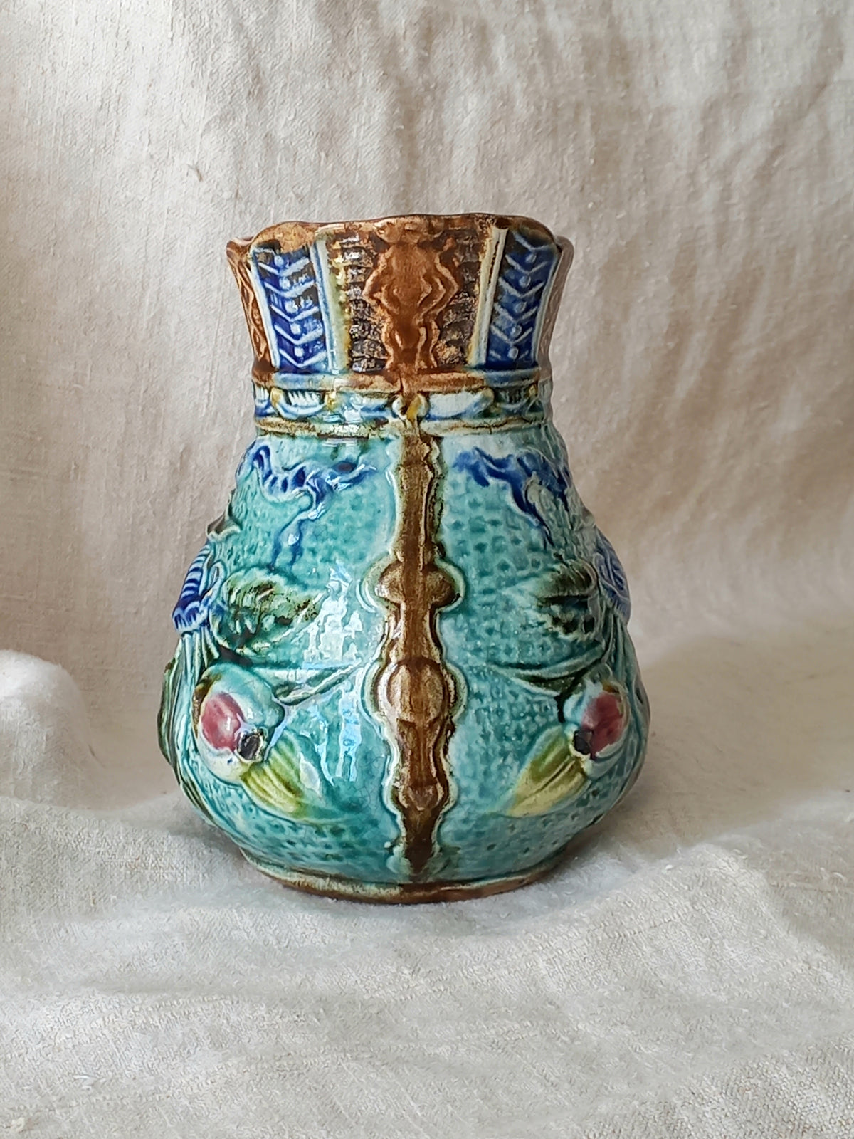 An antique majolica pitcher with a rich glaze set against a draped white fabric background.