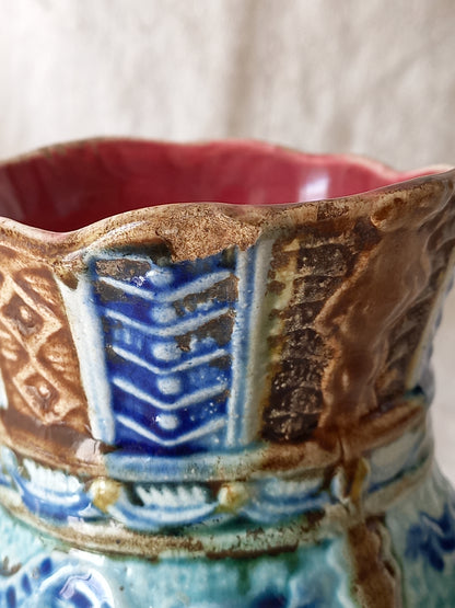 Close-up of an antique majolica pitcher with a rich glaze set against a draped white fabric background.