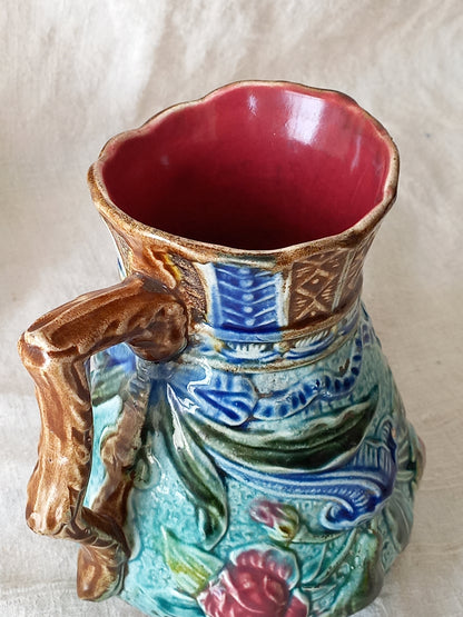 Close-up of an antique majolica pitcher with a rich glaze set against a draped white fabric background.