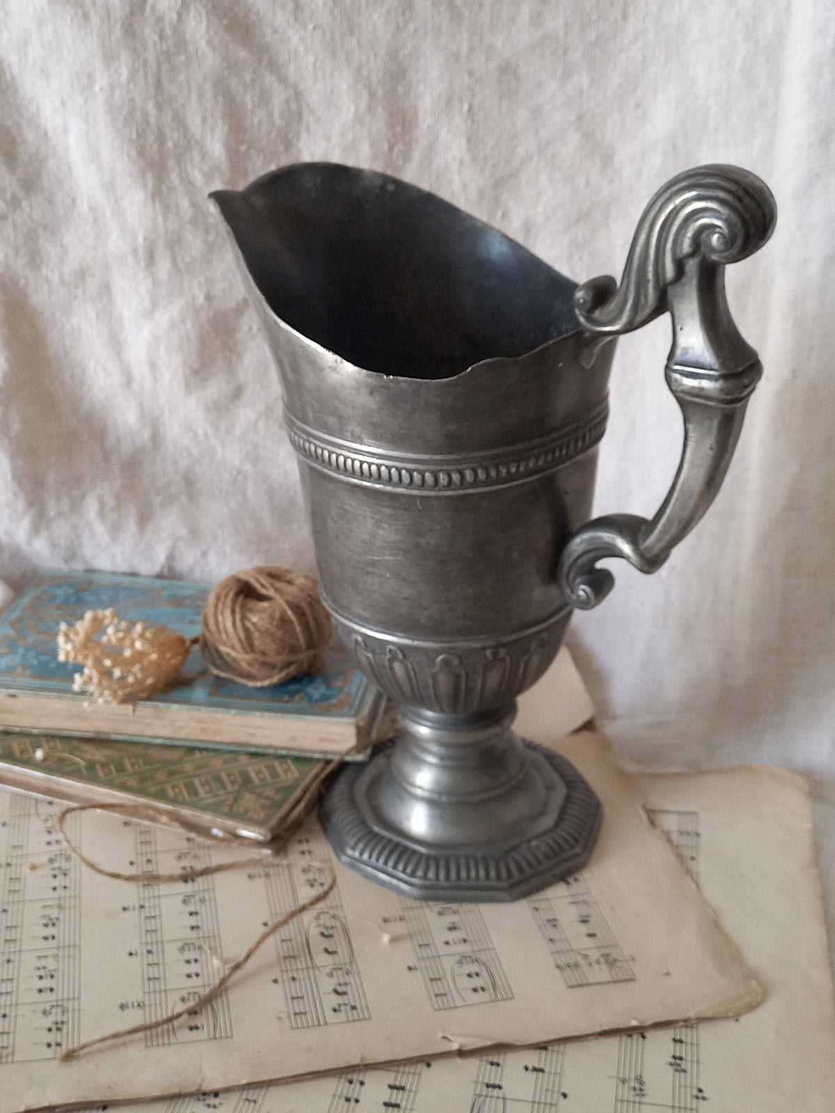 An antique pewter pitcher with a decorative handle, displayed on a rustic background with vintage sheet music, a small book, and twine.