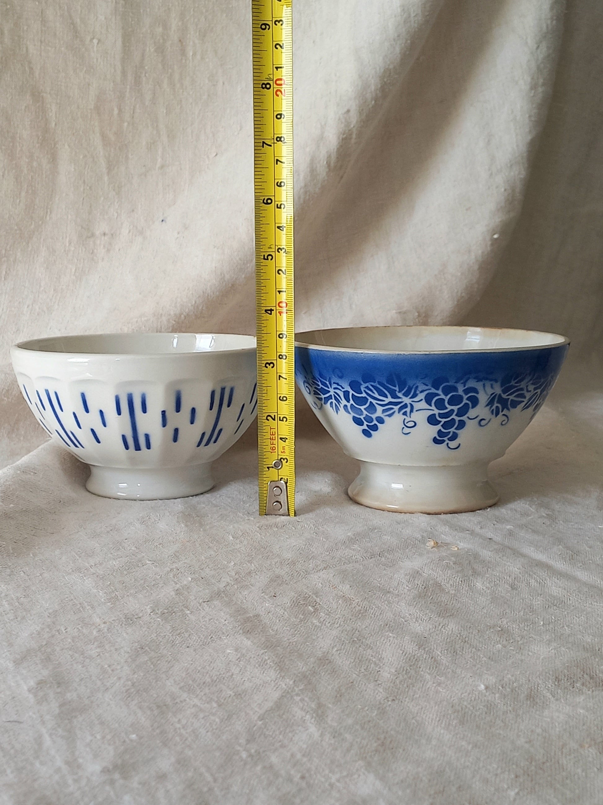 Blue and white vintage ceramic cafe au lait bowls with floral patterns on a light fabric background with a tape measure for reference.