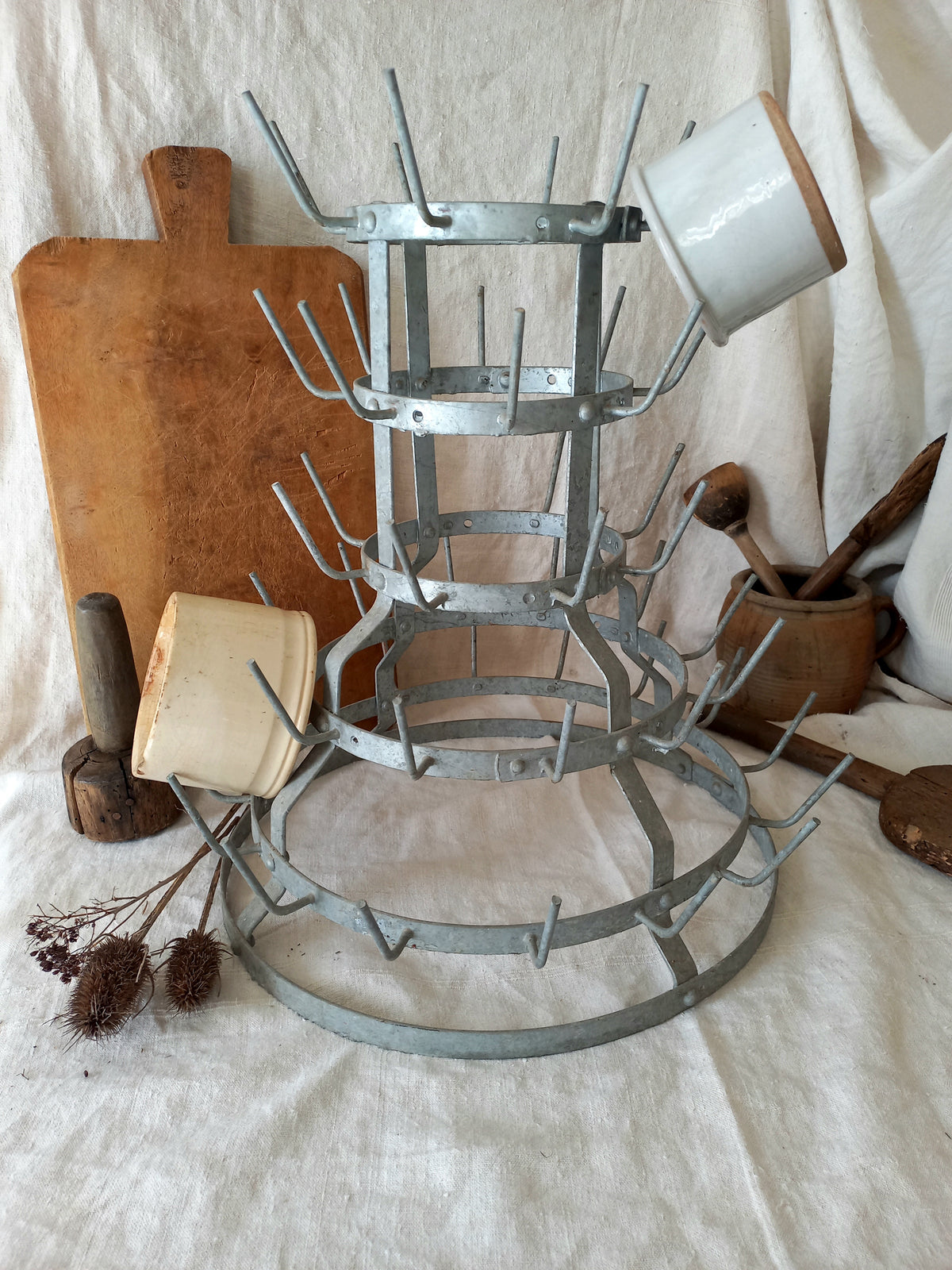A rustic three-tier metal mug rack with wire loops, displayed on a draped white linen cloth with vintage kitchen props including wooden cutting boards, ceramic dishes, and wooden spoons.