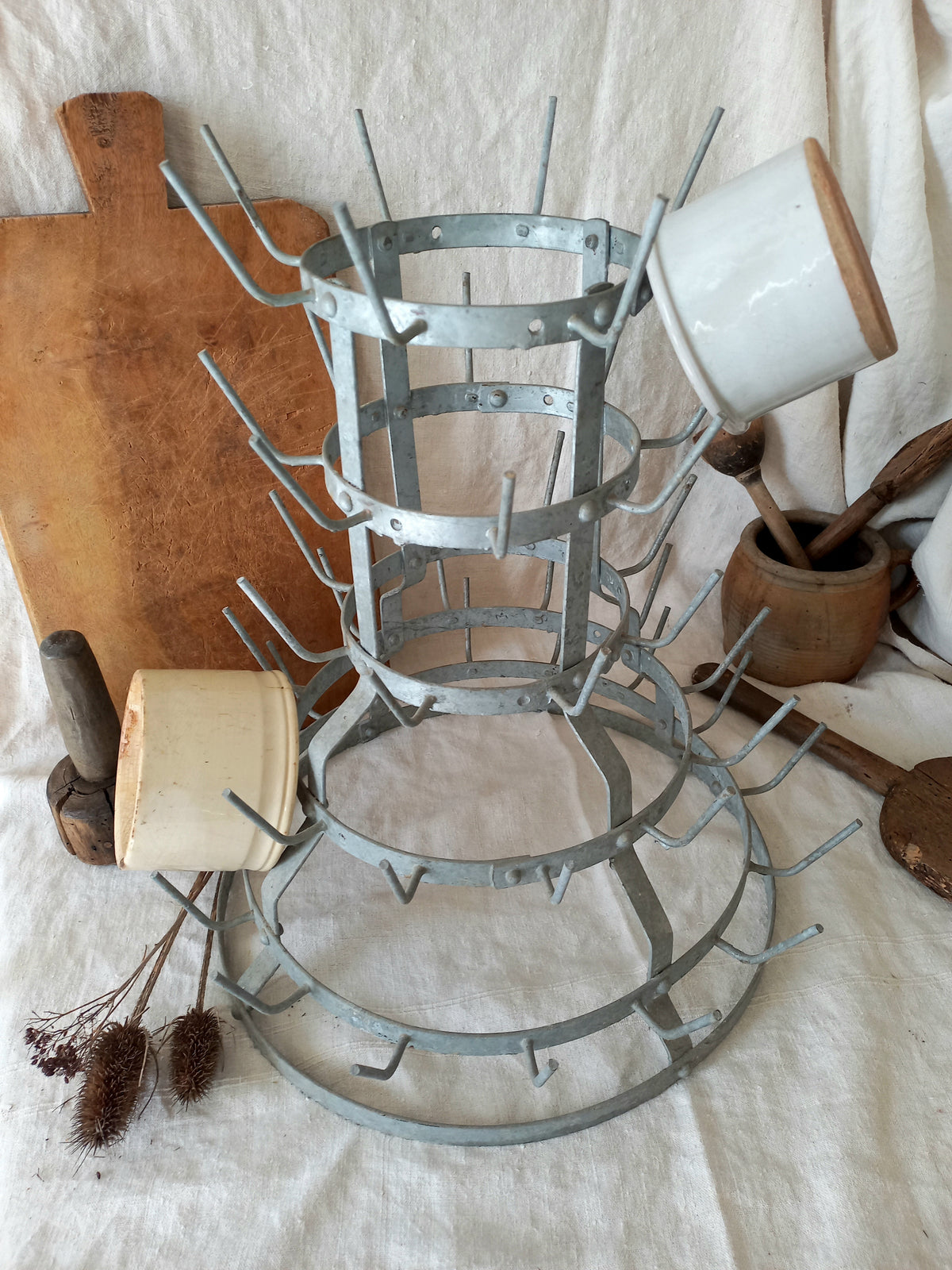 A vintage metal tiered dish rack with multiple hooks, displayed on a white fabric backdrop with rustic kitchen props including a wooden cutting board, ceramic mugs, and wooden utensils.