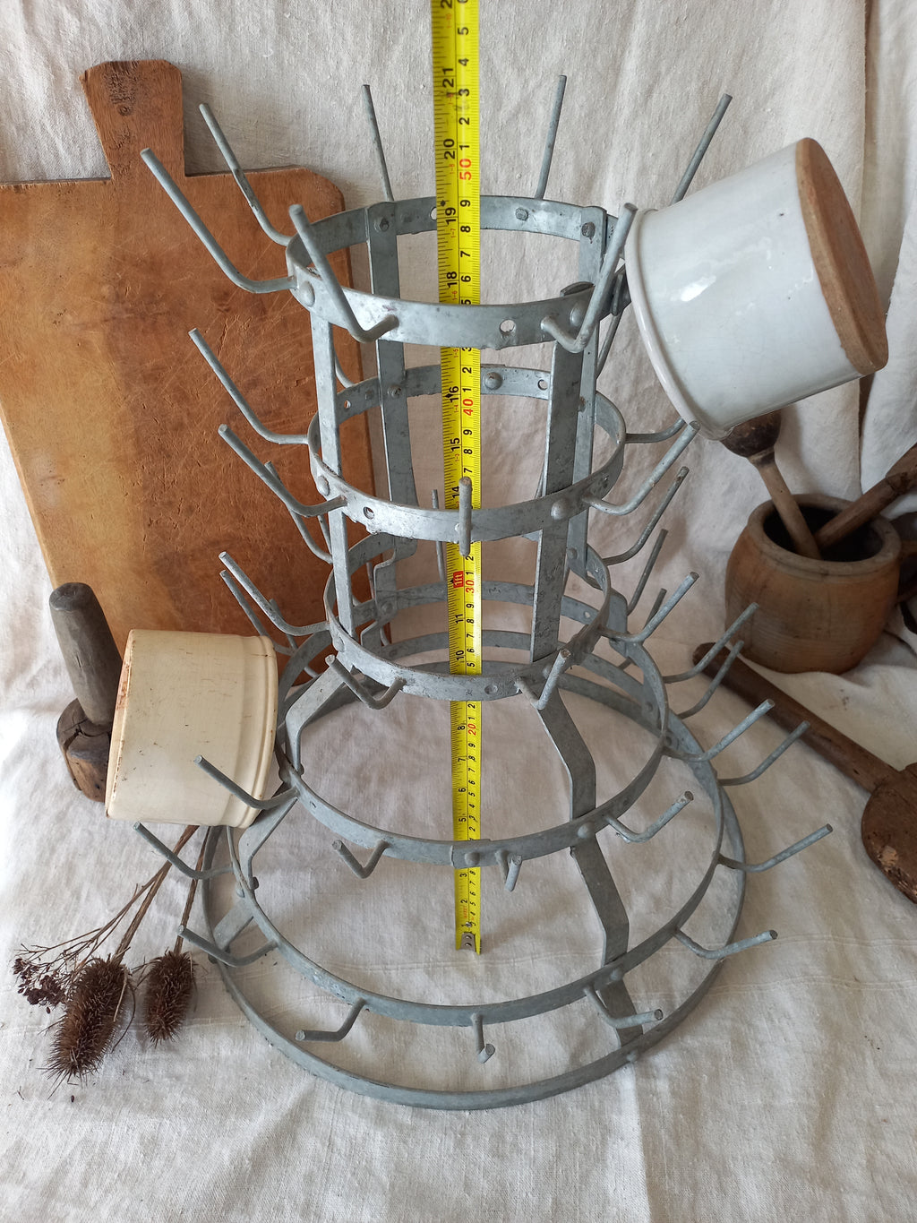 A metal kitchen utensil rack with multiple tiers and hanging hooks, shown standing upright on a white fabric surface. A yellow tape measure is placed vertically through the center of the rack to indicate size.