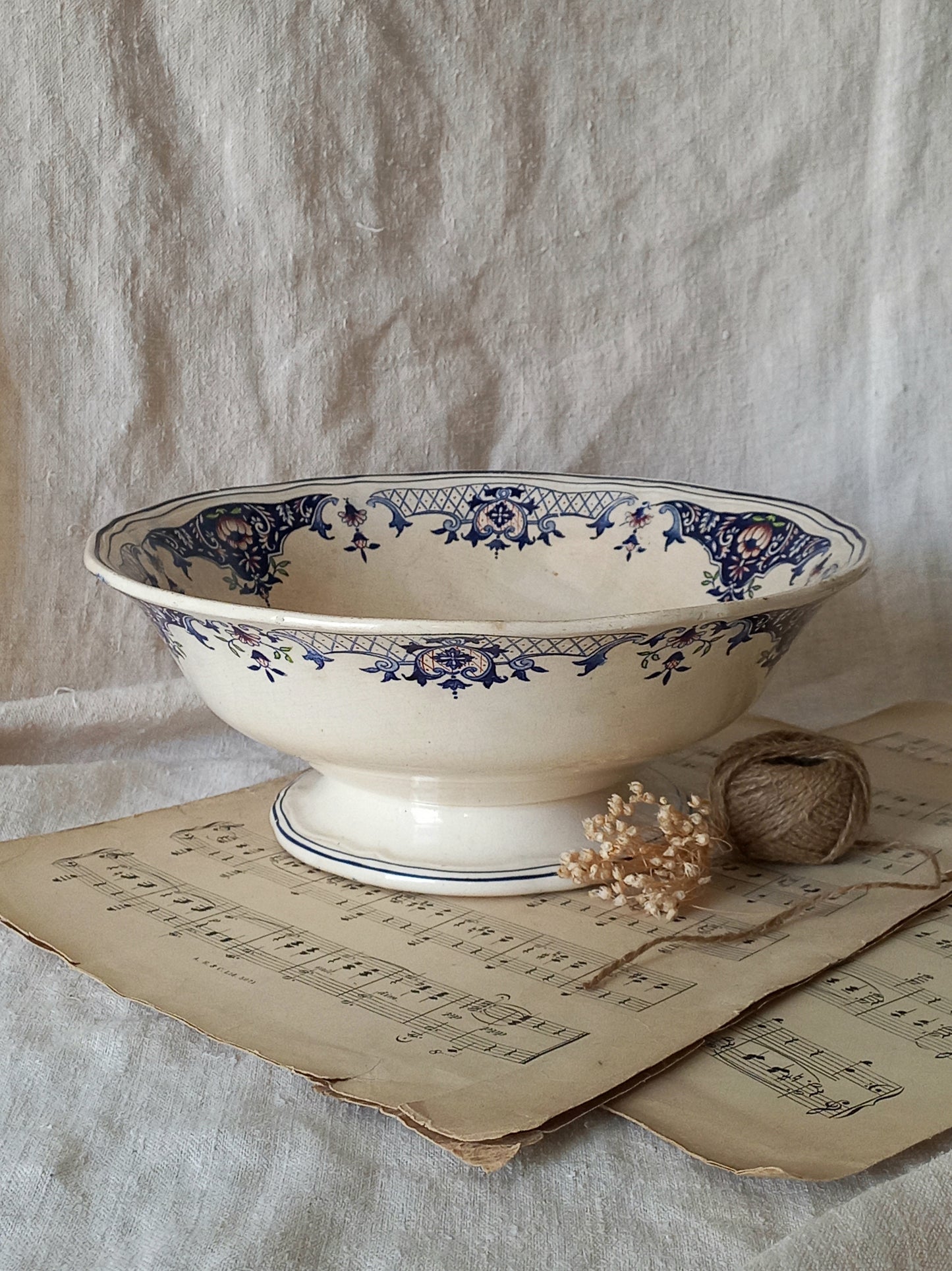 An antique cream-colored ceramic bowl with blue floral patterns, styled on antique sheet music with a small dried flower and twine bundle against a draped linen backdrop.