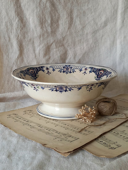 An antique cream-colored ceramic bowl with blue floral patterns, styled on antique sheet music with a small dried flower and twine bundle against a draped linen backdrop.