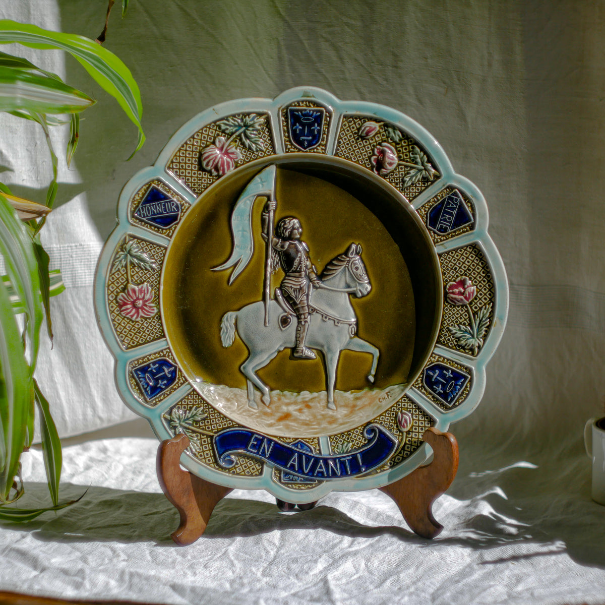 A large vintage/antique majolica wall plate with hand-coloured details in relief, the theme of the plate being the Patron Saint of France - Joan of Arc riding into battle with her banner (in French she is called Jeanne D'Arc).

This wonderful French decorative plate has a rich gloss finish, the glaze even being applied to the underside.

Around the lightly scalloped circumference of this large plate are patriotic symbols such as 'En Avant' (Go Forward), 'Honneur' (Honour) and 'Patrie' (Homeland).