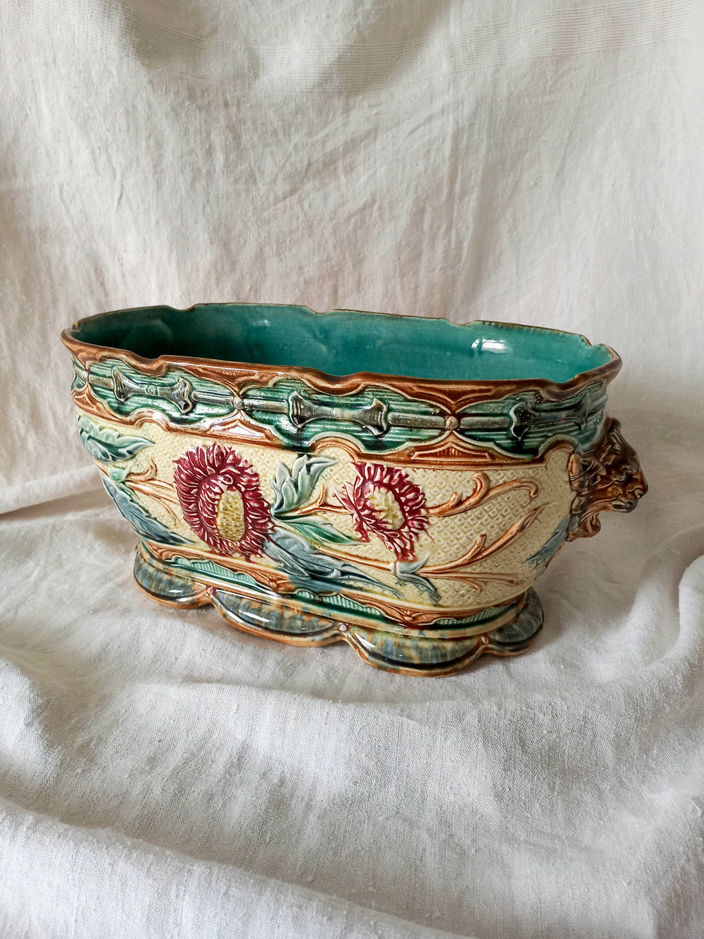 An ornate ceramic planter with a scalloped rim, featuring a turquoise glazed interior and exterior green and gold leaf patterns, decorated with red and yellow floral motifs, set against a draped white fabric background.