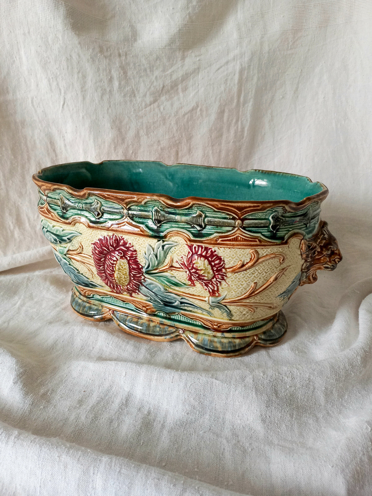 An ornate ceramic planter with a scalloped rim, featuring a turquoise glazed interior and exterior green and gold leaf patterns, decorated with red and yellow floral motifs, set against a draped white fabric background.