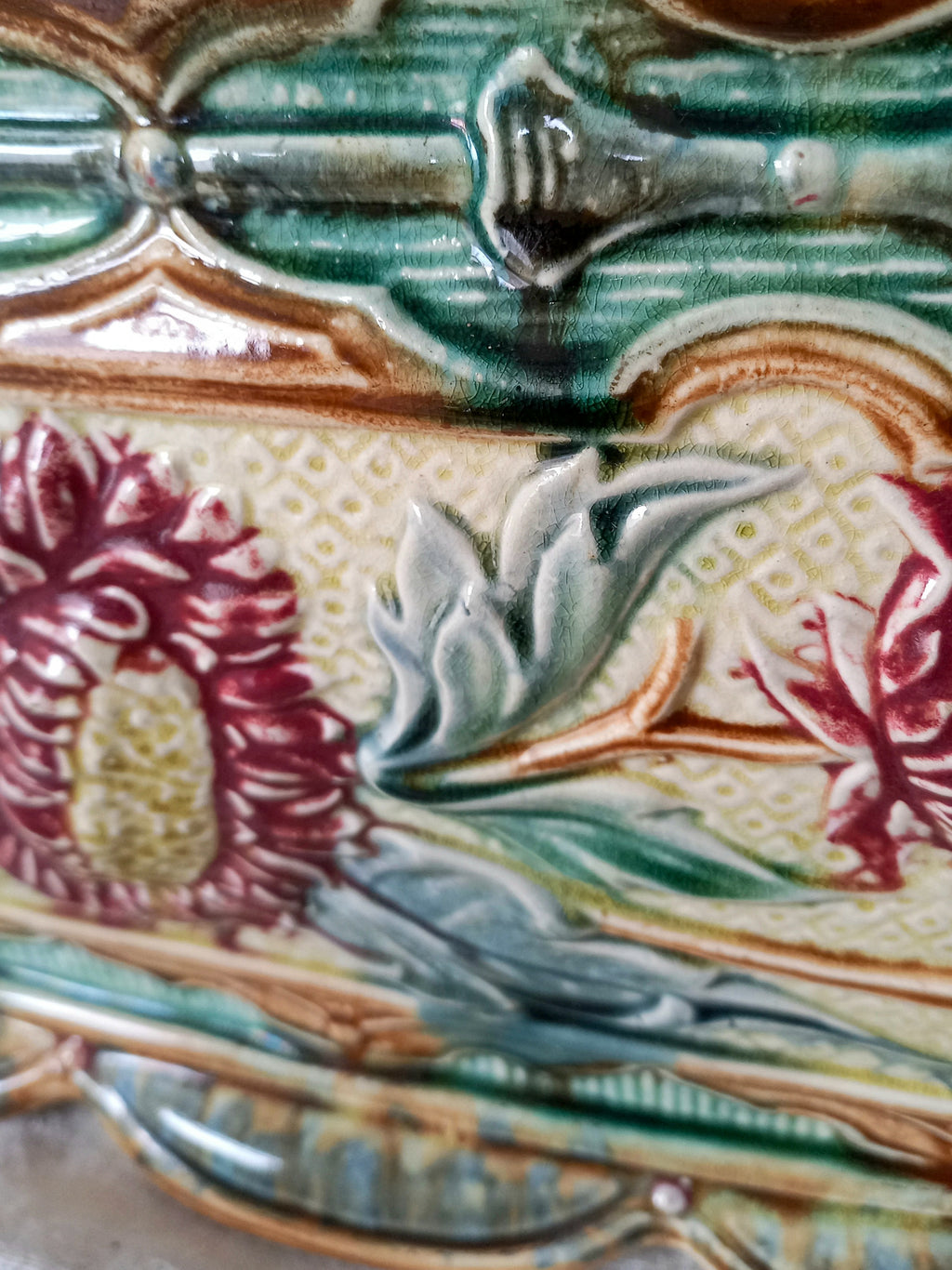 Close-up of a decorative ceramic surface with raised floral and scroll motifs in glossy green, cream, burgundy red, and tan glaze.