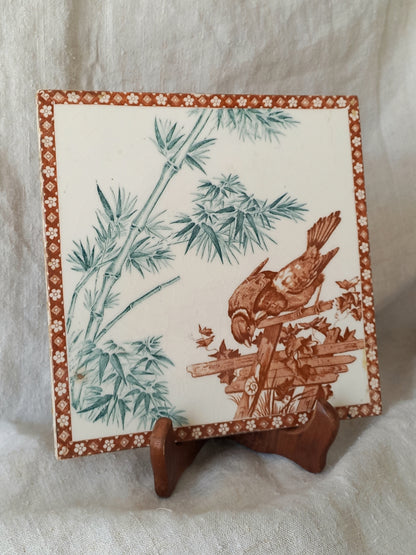 A square ceramic tile with a brown bird perched on a branch among green bamboo leaves, displayed on a small wooden stand against a white fabric background.