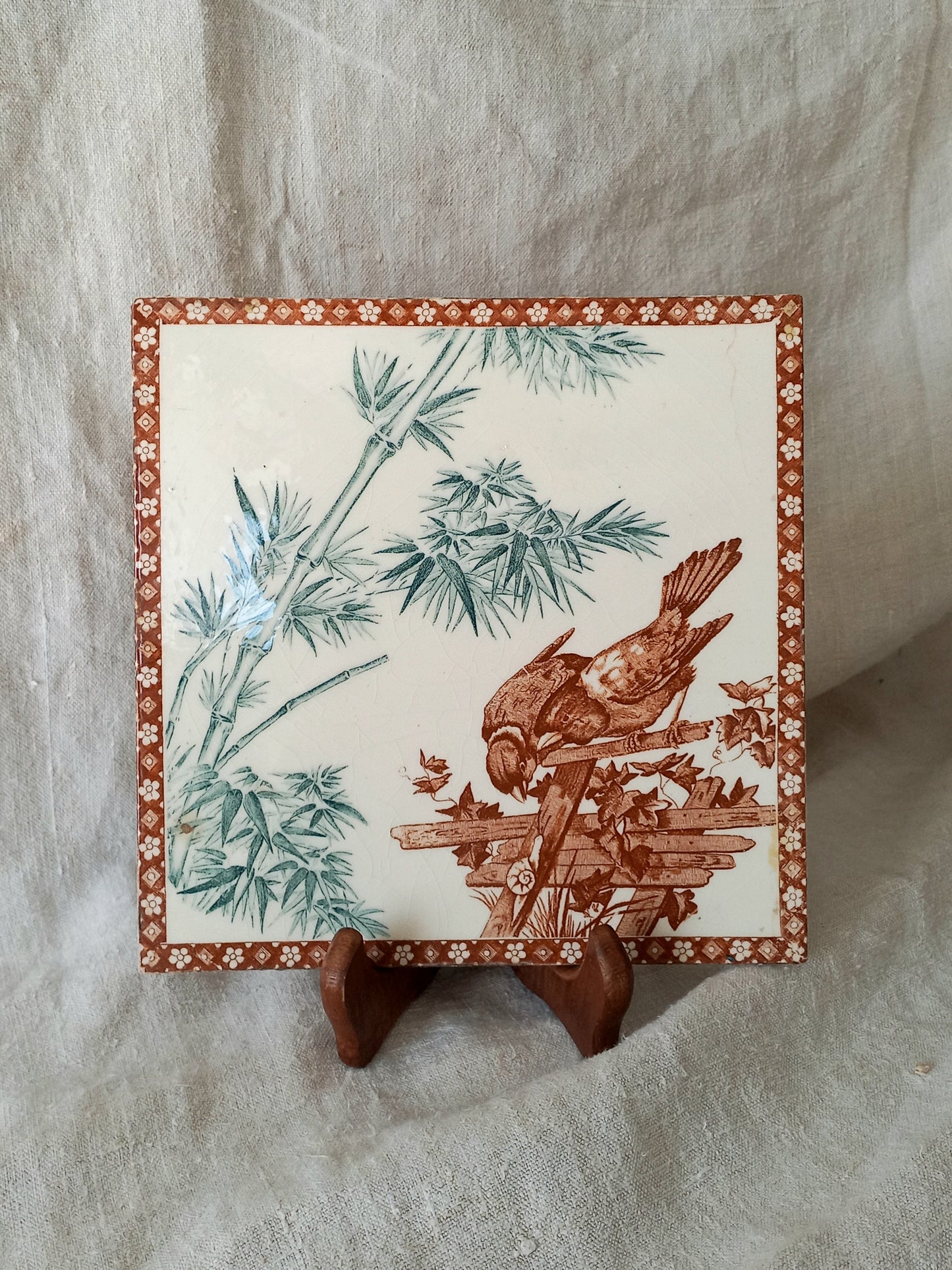 A square ceramic tile with a brown bird perched on a branch among green bamboo leaves, displayed on a small wooden stand against a draped beige fabric background.