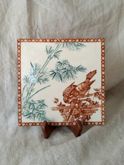 A square ceramic tile with a brown bird perched on a branch among green bamboo leaves, displayed on a small wooden stand against a draped beige fabric background.