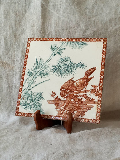 A square ceramic decorative tile with a brown and green bamboo and bird print on an off-white background, displayed on a small wooden stand against a textured beige fabric backdrop.