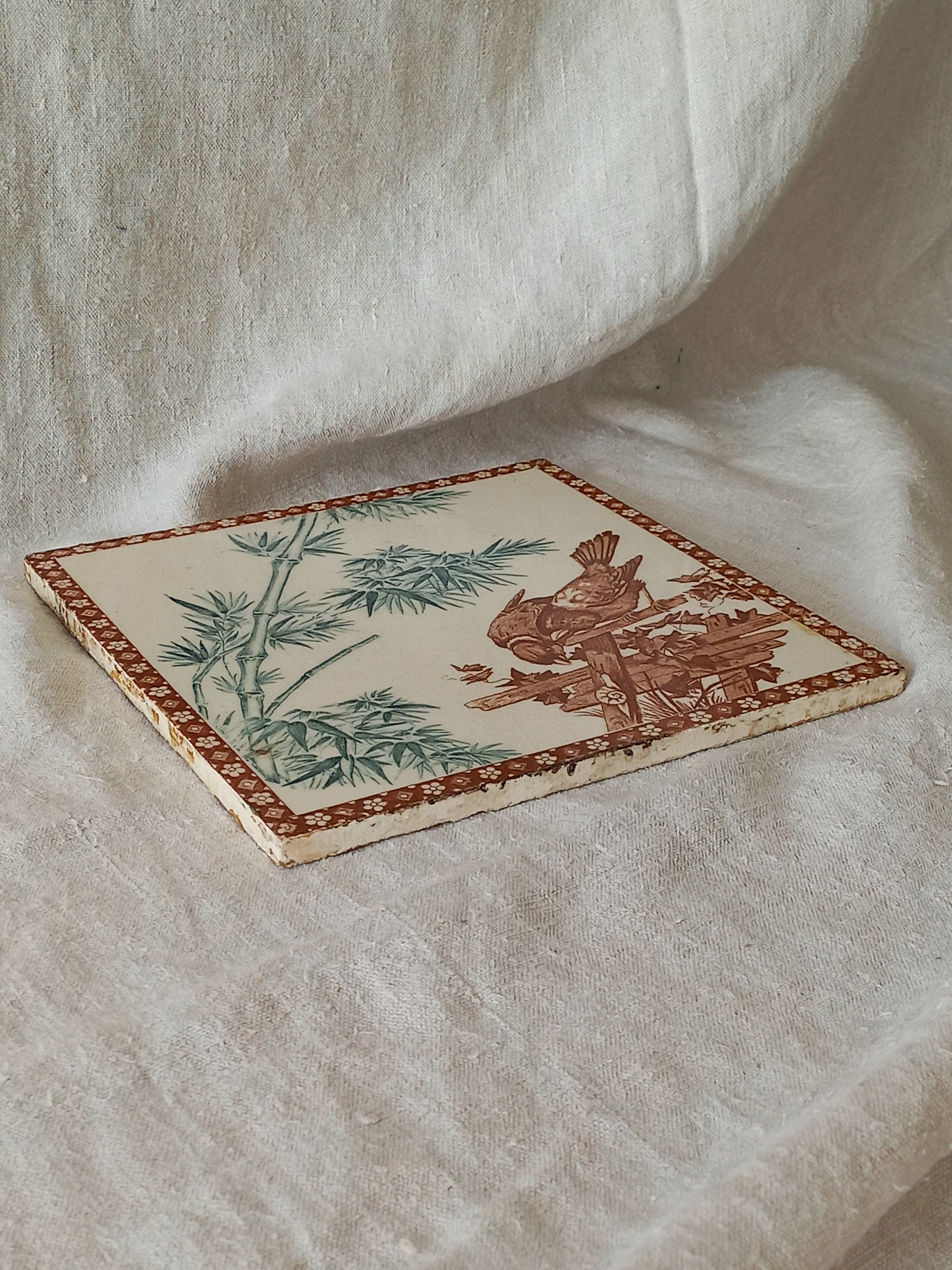 A square ceramic tile featuring a traditional style illustration of a bird and bamboo in muted green and brown tones on an off-white background, bordered by a brown patterned edge, displayed on draped cream fabric.