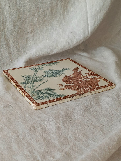 A square ceramic tile featuring a traditional style illustration of a bird and bamboo in muted green and brown tones on an off-white background, bordered by a brown patterned edge, displayed on draped cream fabric.