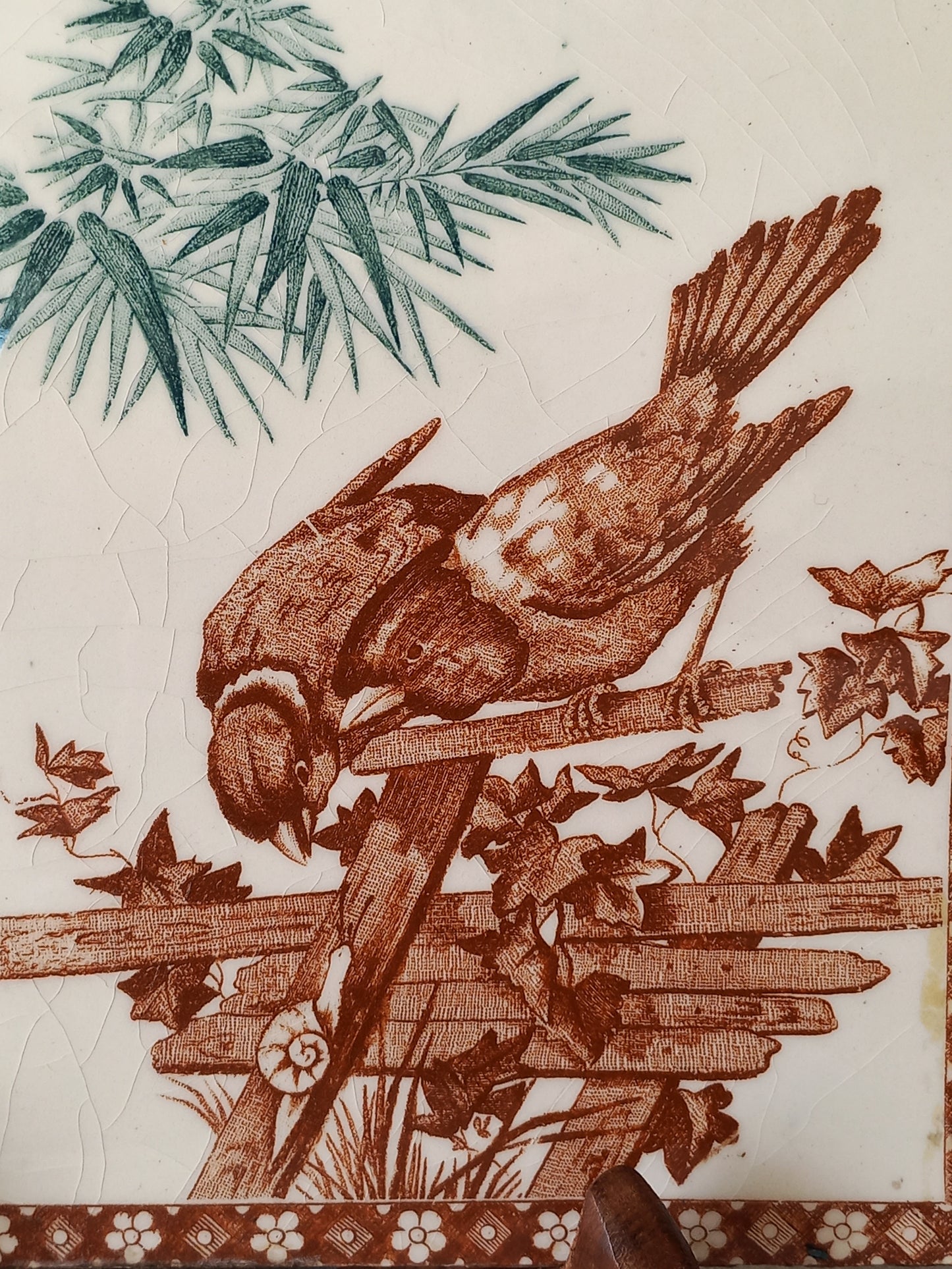 A close-up of an antique tile featuring a brown bird perched on a fence with leaves, alongside green foliage on the left and a patterned brown border on the right, on an off-white background.