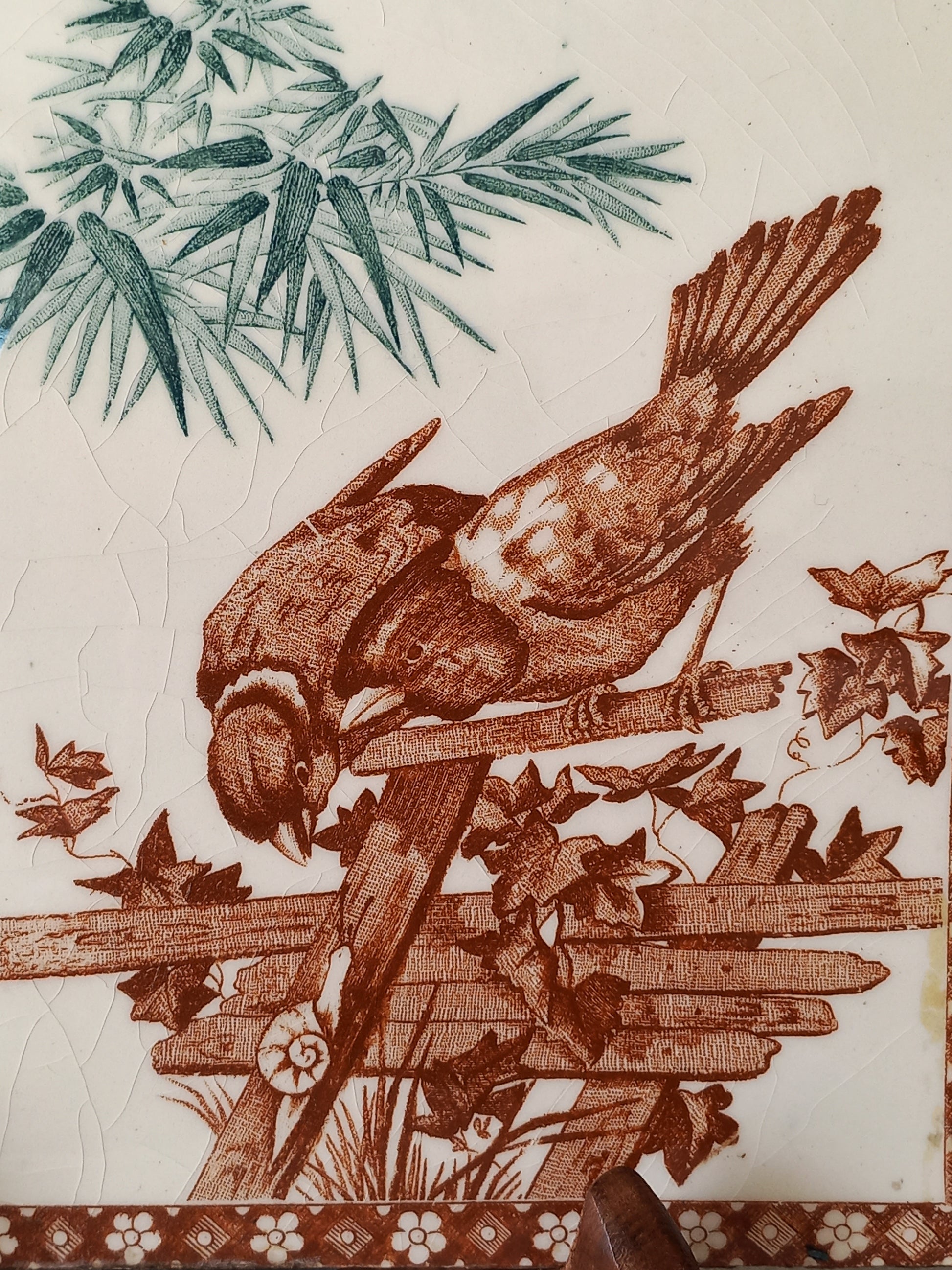 A close-up of an antique tile featuring a brown bird perched on a fence with leaves, alongside green foliage on the left and a patterned brown border on the right, on an off-white background.