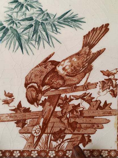 A close-up of an antique tile featuring a brown bird perched on a fence with leaves, alongside green foliage on the left and a patterned brown border on the right, on an off-white background.