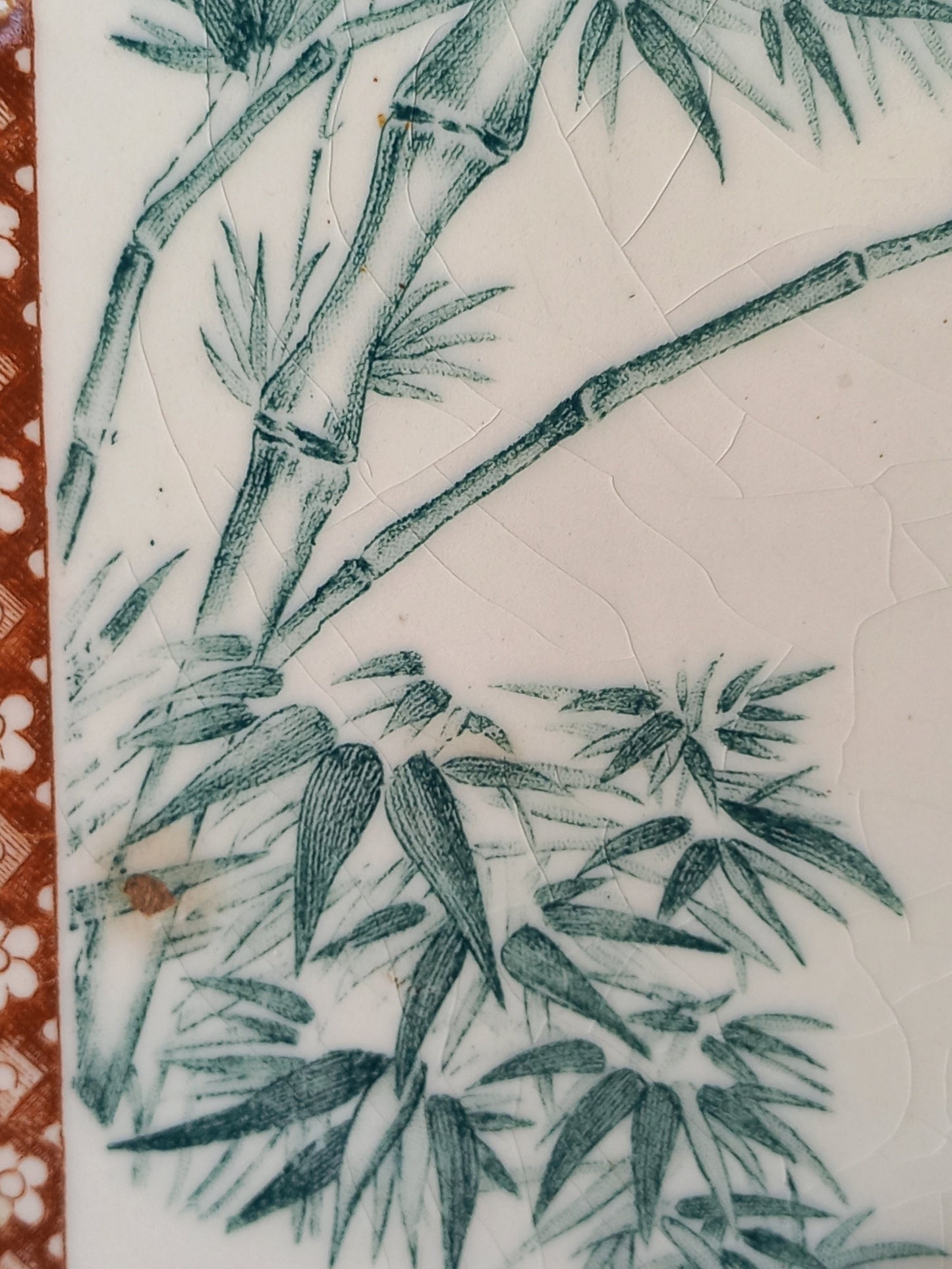 Close-up of a white ceramic tile or plaque featuring a green hand-painted bamboo design, with a textured crackled glaze and a decorative reddish-brown patterned border on the left edge.