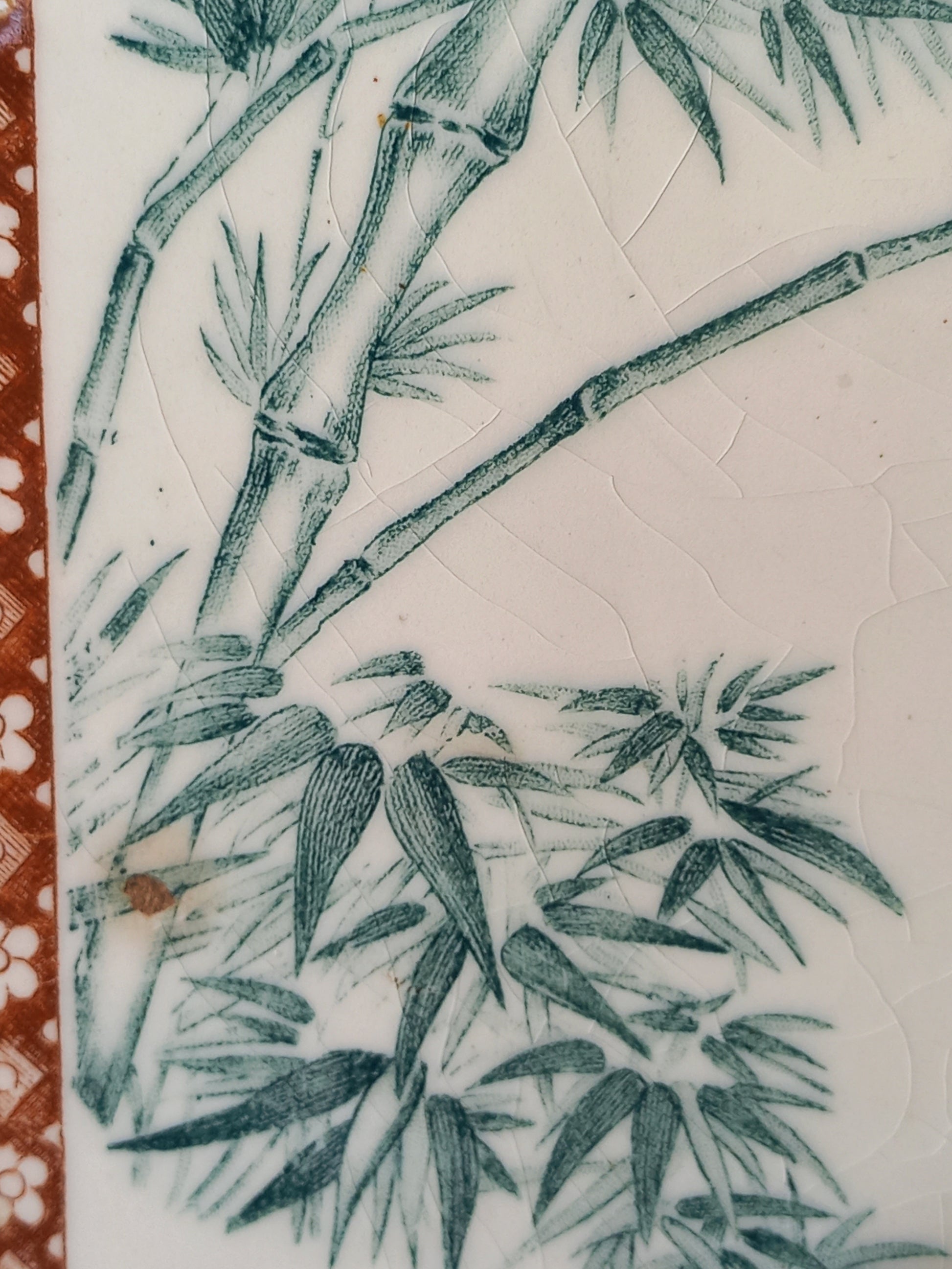 Close-up of a white ceramic tile or plaque featuring a green hand-painted bamboo design, with a textured crackled glaze and a decorative reddish-brown patterned border on the left edge.