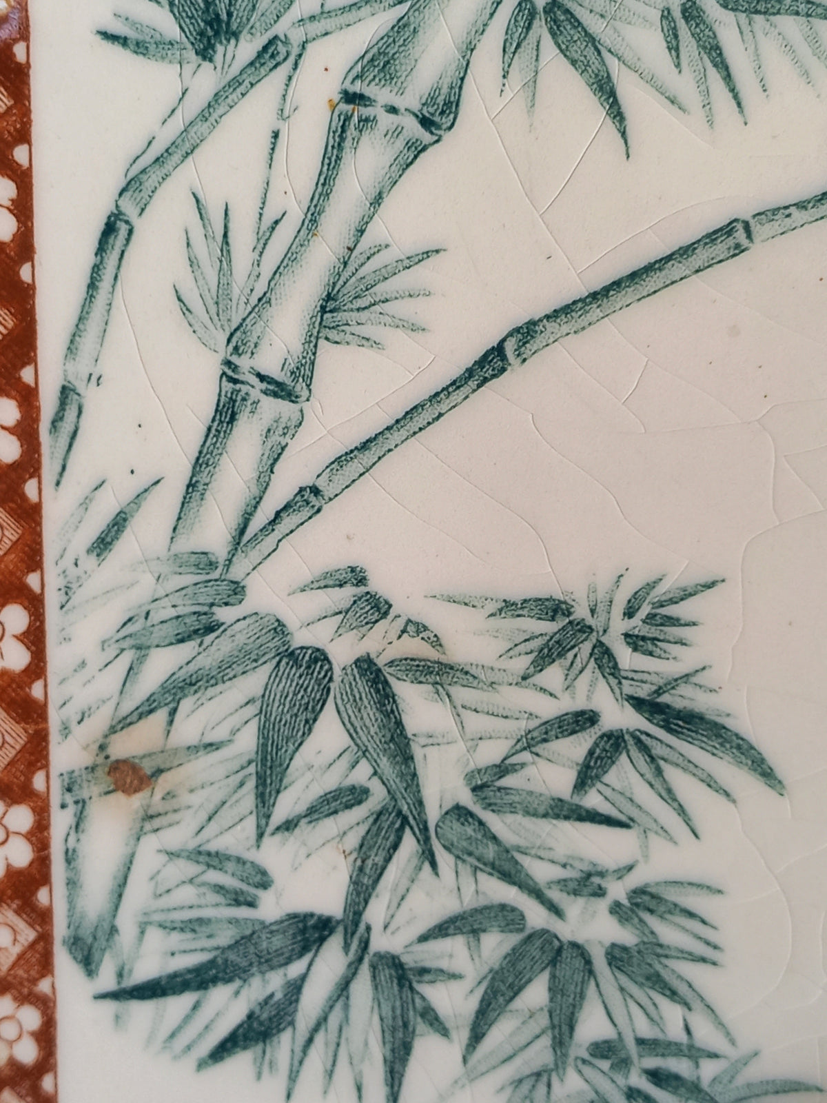 Close-up of a white ceramic tile or plaque featuring a green hand-painted bamboo design, with a textured crackled glaze and a decorative reddish-brown patterned border on the left edge.