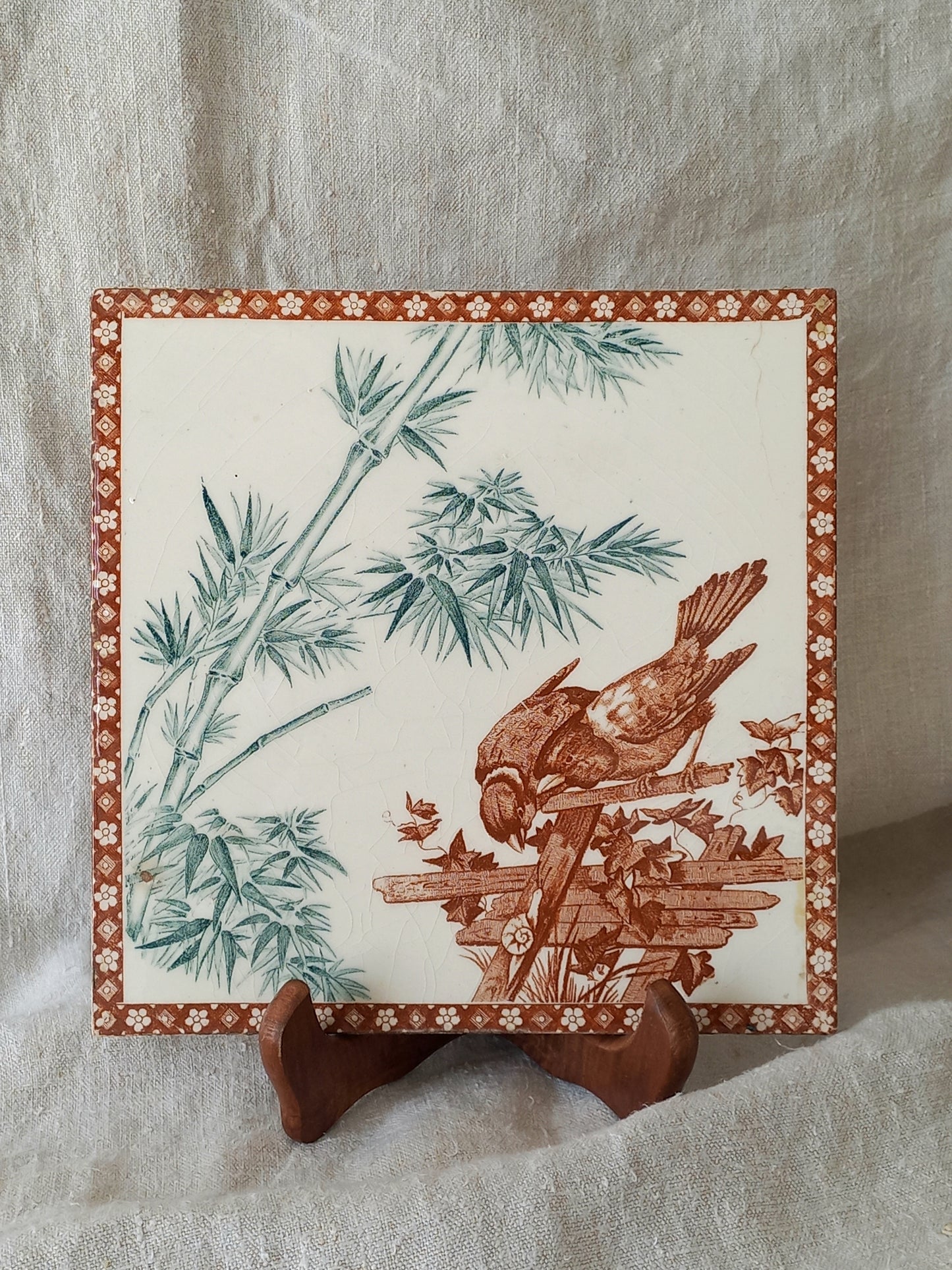 A square ceramic tile with a brown and green bamboo and bird design on an off-white background, displayed on a small wooden stand against a draped beige fabric backdrop.