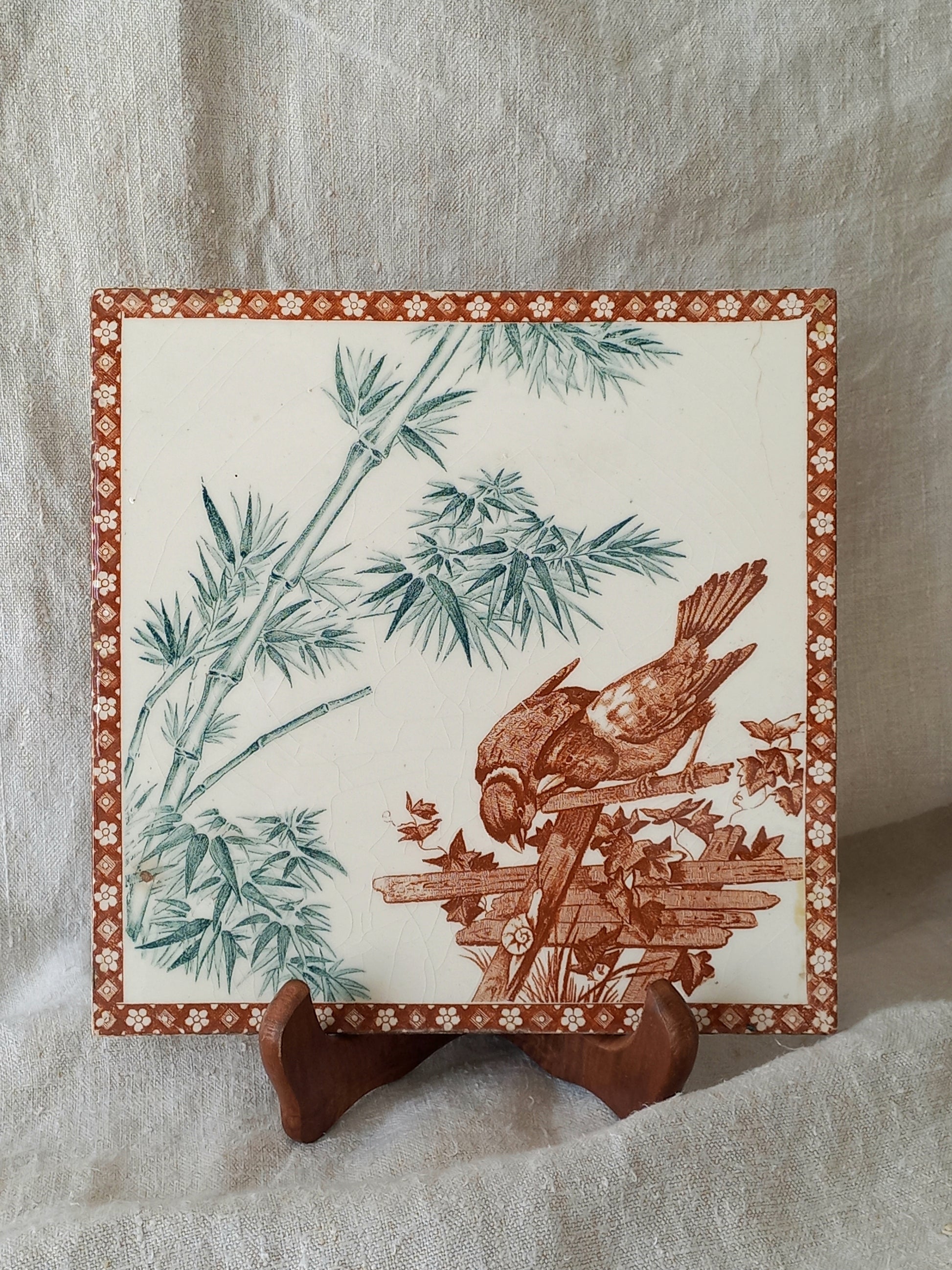 A square ceramic tile with a brown and green bamboo and bird design on an off-white background, displayed on a small wooden stand against a draped beige fabric backdrop.