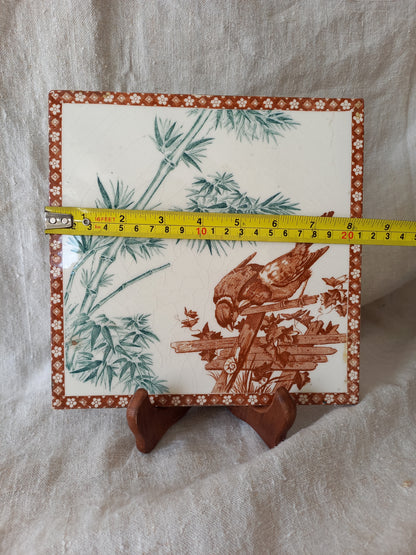 A square ceramic tile with a brown and green  illustration of a bird on a wooden structure, bordered by a floral pattern, shown on a white fabric background with a yellow tape measure for scale.