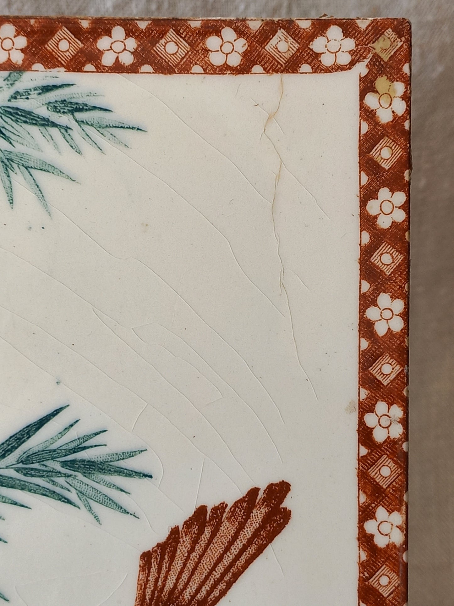 Close-up of a decorative ceramic tile or plate corner featuring a white center, a rust-brown patterned border with white floral motifs, and partial green botanical illustrations.