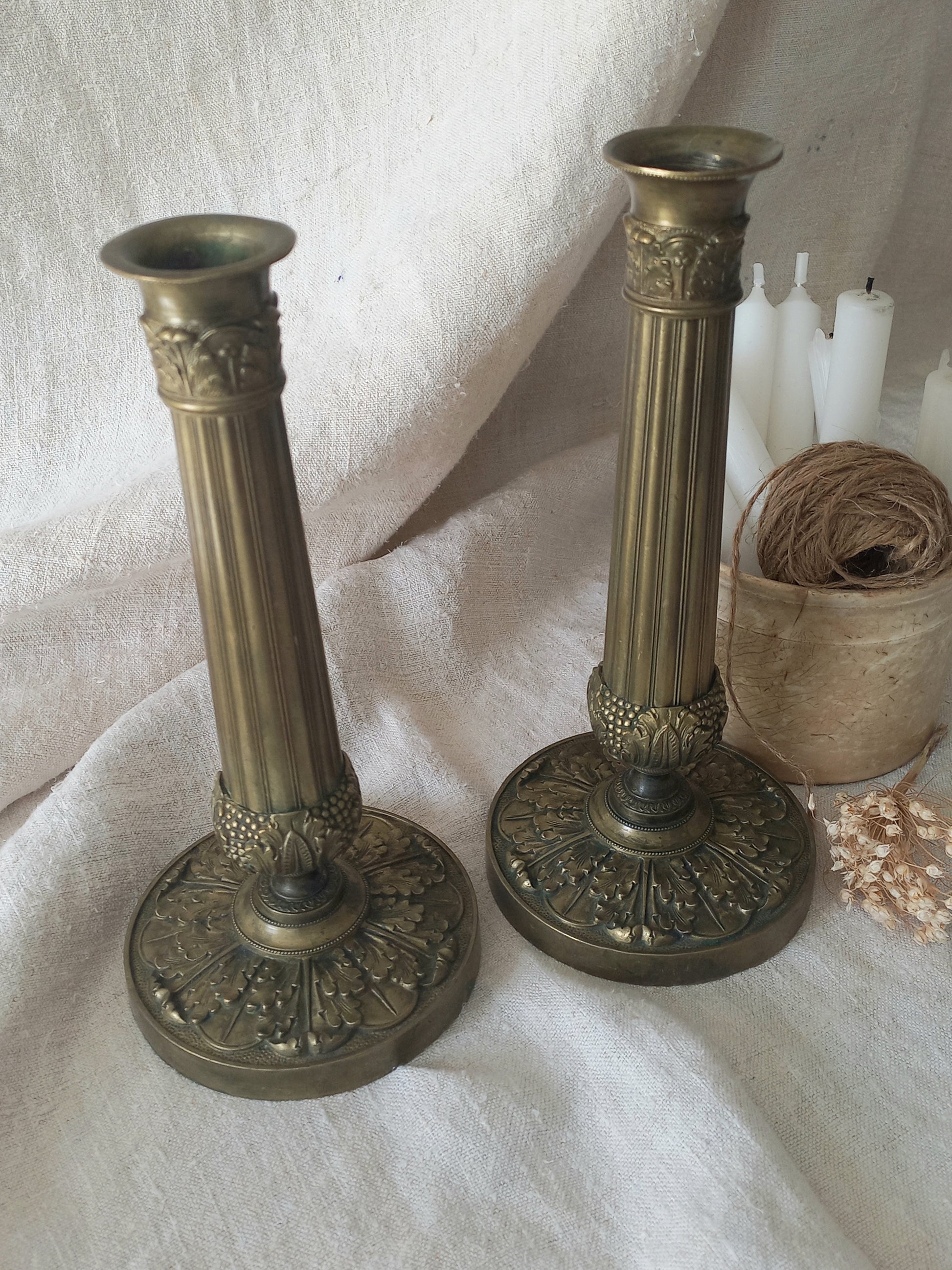 Two ornate brass candlesticks on a textured white surface with twine and dried flowers.