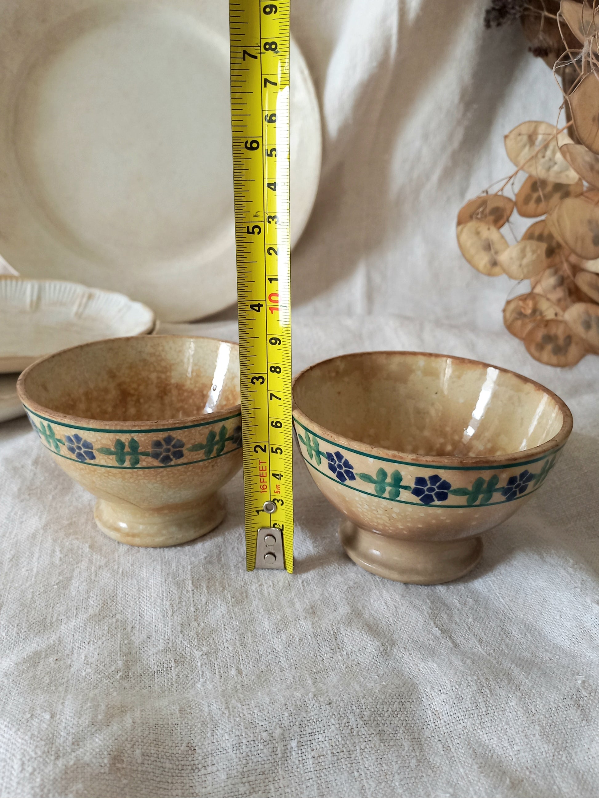 Two ceramic bowls with floral patterns on a textured surface, measured by a tape measure.