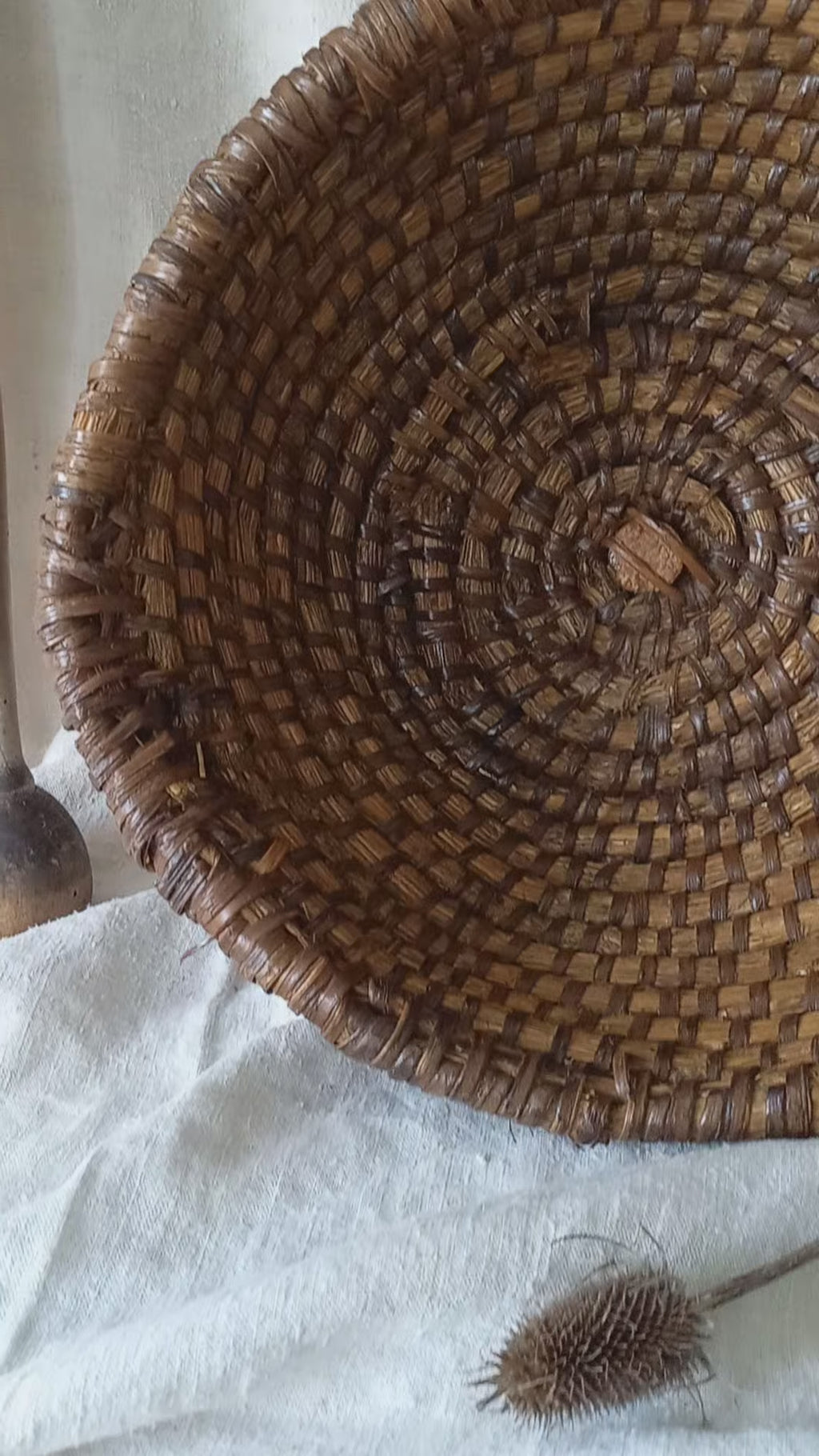 A large round woven vintage basket made of dark brown natural fibers, styled on a draped white linen cloth with rustic wooden kitchen utensils in the background.