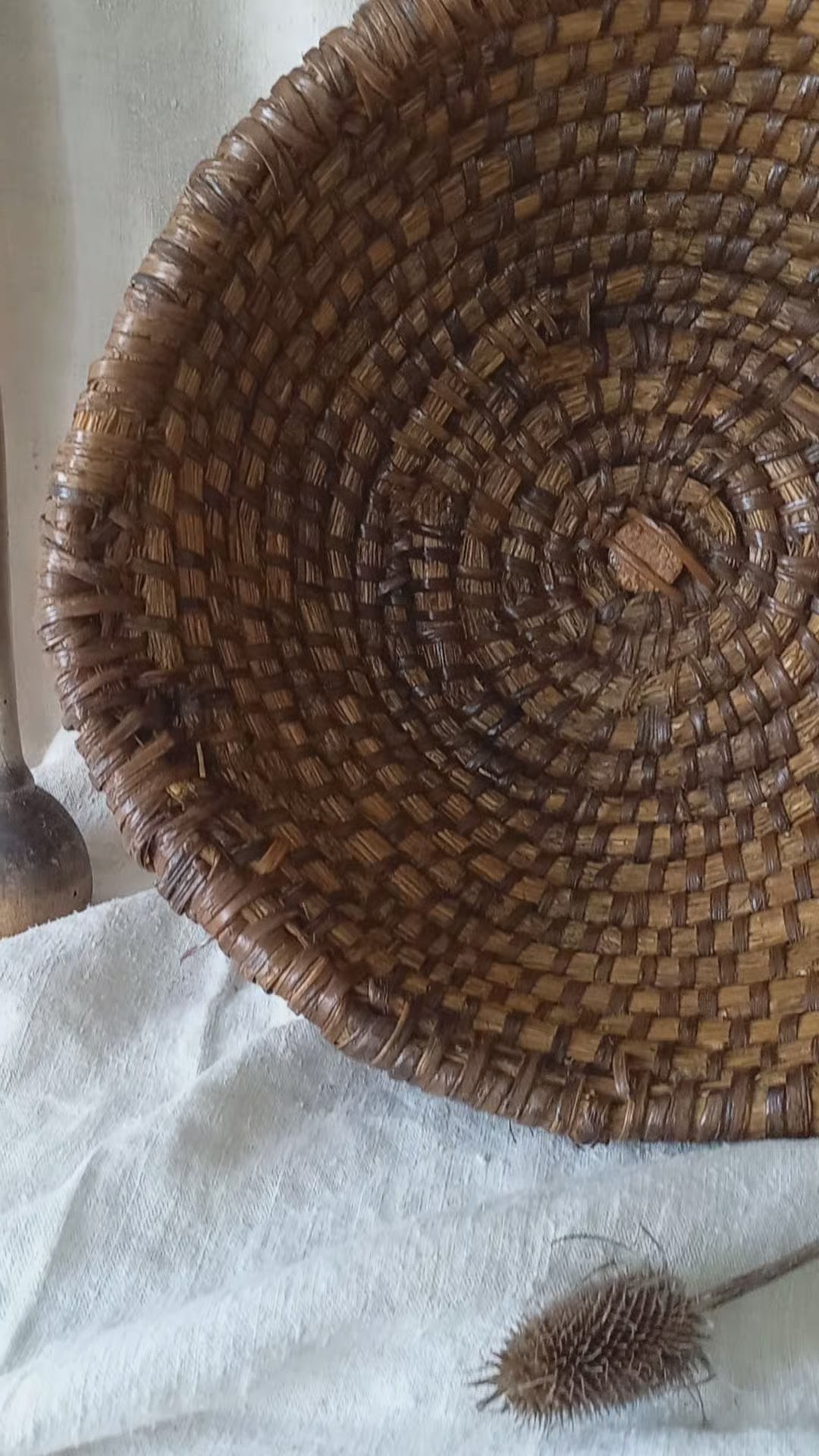 A large round woven vintage basket made of dark brown natural fibers, styled on a draped white linen cloth with rustic wooden kitchen utensils in the background.