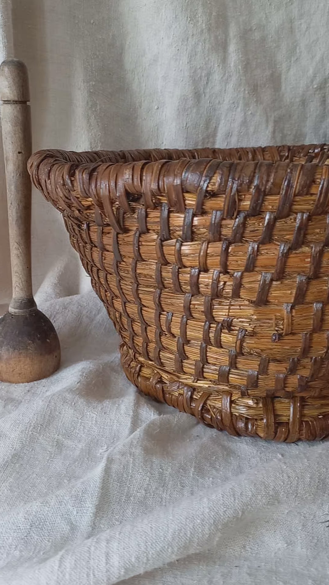 A large vintage round woven vintage basket made of dark brown natural fibers, styled on a draped white linen cloth with rustic wooden kitchen utensils in the background.