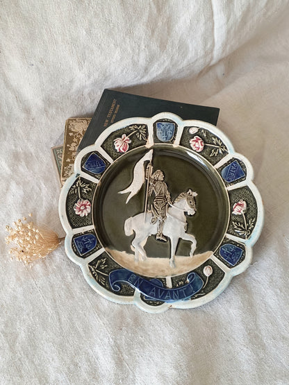 Decorative plate with a scene of Joan of Arc on a horse, surrounded by decorative elements on a white background.