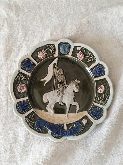 Decorative plate with a scene of Joan of Arc on a horse, surrounded by decorative elements on a white background.
