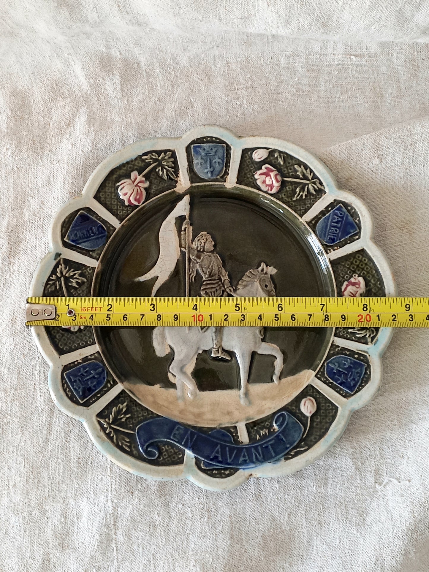 Decorative plate with a scene of Joan of Arc on a horse, surrounded by decorative elements on a white background and a tape measure for reference.
