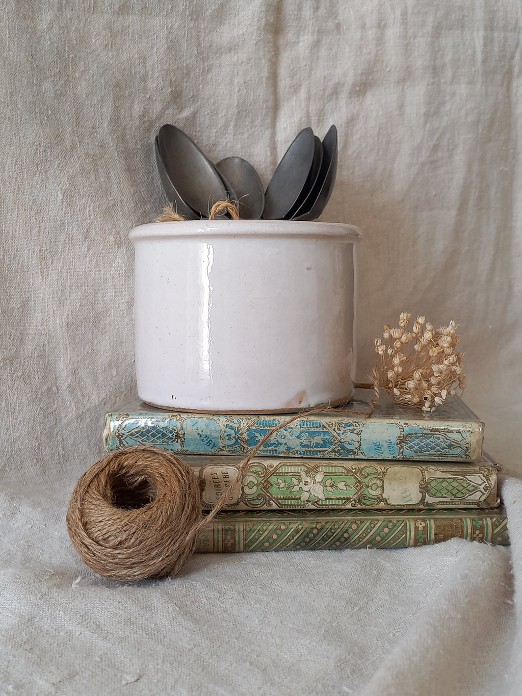 Antique jam pot on a textured surface containing spoons, dried flowers, and books on a textured surface